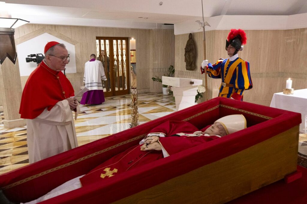 El papa descansa sobre un féretro de madera forrado de terciopelo rojo con una casulla purpura y una mitra blanca y en las manos lleva un rosario.
